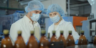 Two specialists, a young man and a middle-aged woman, in white coats, disposable hats and medical masks, are discussing production issues against the backdrop of plastic bottles.